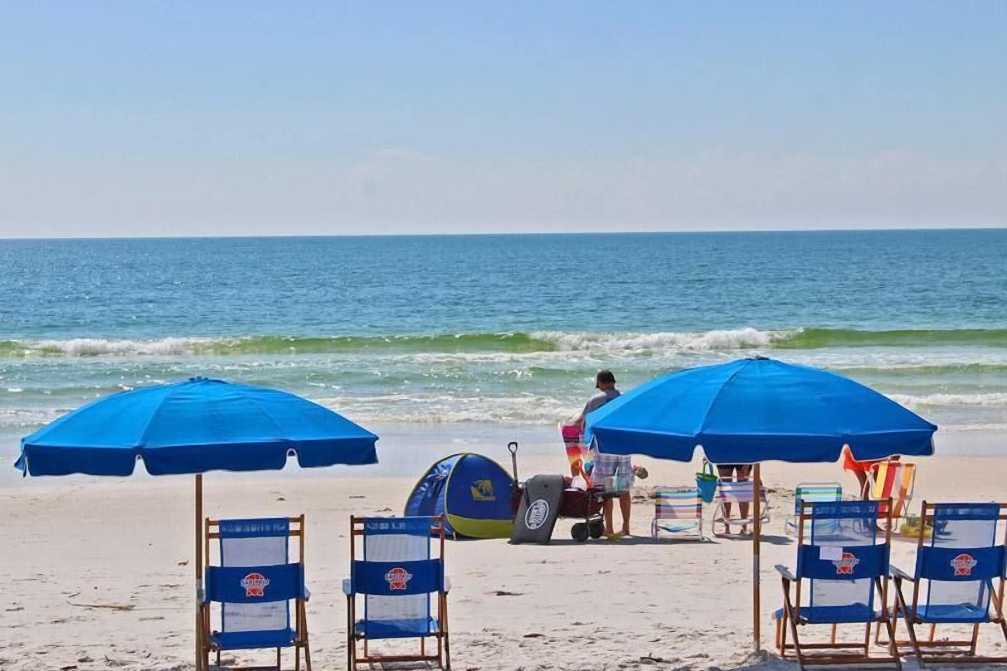 A beach with blue umbrellas, beach chairs, and a person with a surfboard.