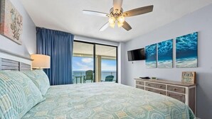 A bedroom with a bed, a ceiling fan, a TV, and a view of the ocean.