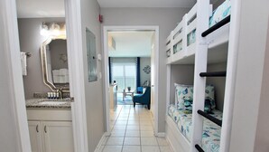 Un couloir étroit avec une salle de bain sur la gauche, un coin salon avec une table et des chaises au centre, et des lits superposés sur la droite.