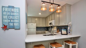 A kitchen with white cabinets, a white countertop, and three wicker stools. There is a sign on the wall that reads "FRIENDS ARE LIKE SEASHELLS. WE COLLECT THEM ALONG THE WAY."