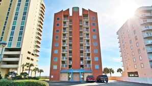 A multi-story hotel with a central entrance, parking area, and adjacent buildings. - Westwind 301 (Gulf Shores)
