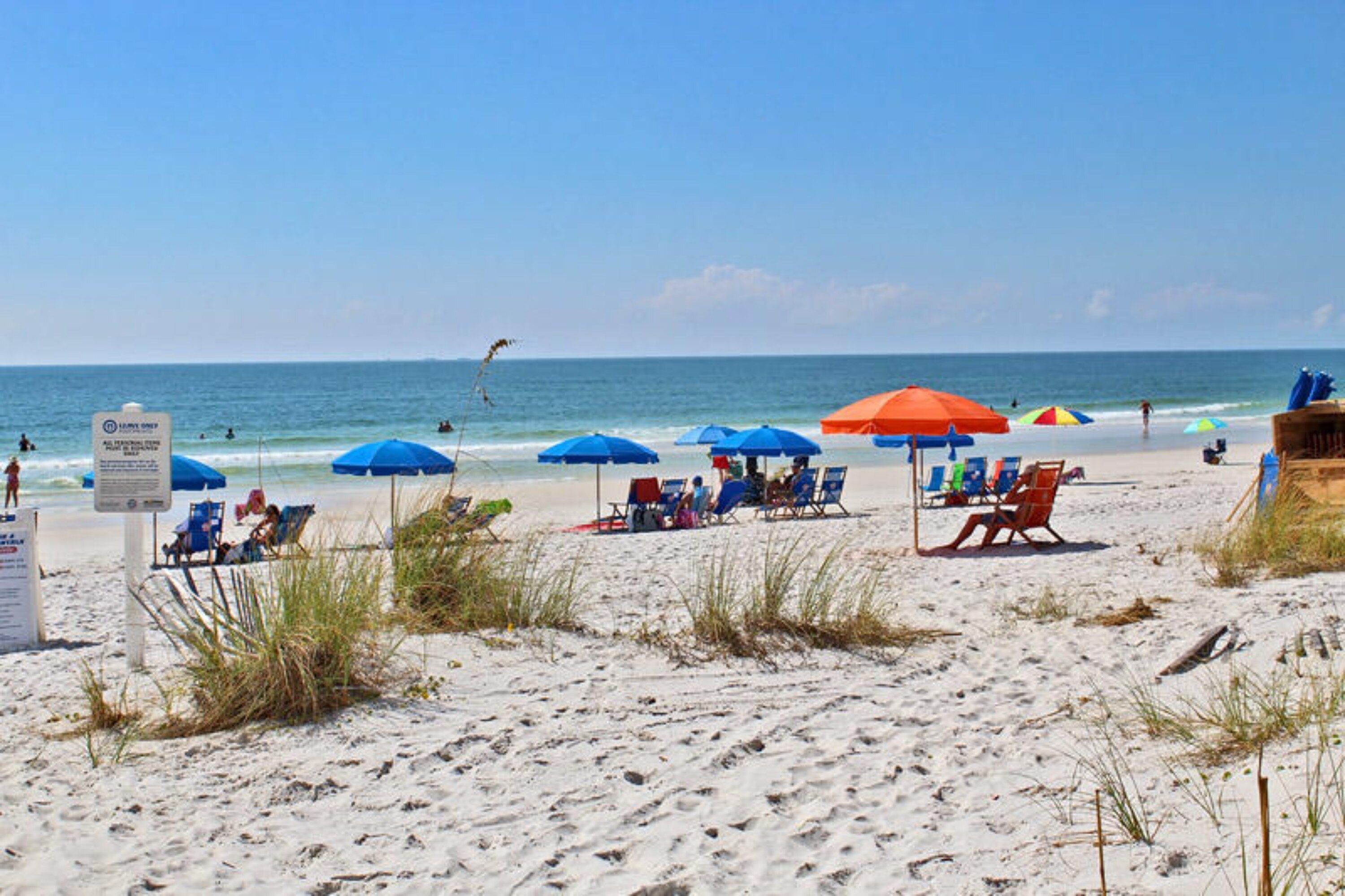 Ein Strand mit mehreren blauen und orangefarbenen Sonnenschirmen, Liegestühlen und einem Schild, das eine Rettungsschwimmerstation kennzeichnet.