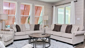 Ein Wohnzimmer mit einem beigefarbenen Sofa, einem runden Couchtisch und zwei Sessel. Große Fenster mit Holzjalousien bieten Blick auf die grüne Umgebung.