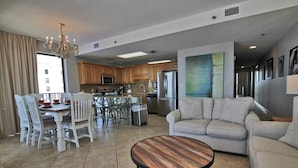 A spacious living room with a round wooden coffee table, a beige sofa, and a dining area with white chairs and a table. - Phoenix East 1201 (Orange Beach)