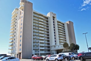 A multi-story hotel with a central tower, a parking lot with several cars, and a clear blue sky. - Summerchase 1108 (Orange Beach)