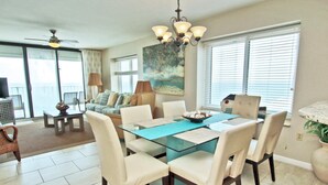 A dining area with a glass table, white chairs, and a chandelier. There is a sofa and a painting on the wall. - Summerchase 1108 (Orange Beach)