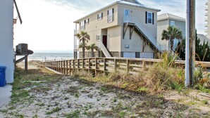 A beachfront house with a wooden deck, a staircase leading to a balcony, and a view of the ocean.
