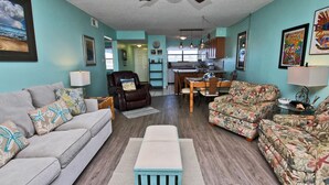 A living room with a sofa, armchair, and patterned armchair, a dining table with chairs, a kitchen area, and a ceiling fan.