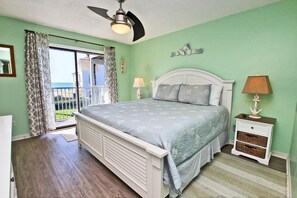 Un dormitorio con cama, mesita de noche, lámpara, ventilador de techo, ventana con cortinas y vista al océano.