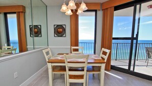 Uma área de refeições com mesa e cadeiras, janelas amplas com vista para o oceano e uma varanda com guarda-corpo.