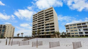 Un hotel frente a la playa con varios pisos, una playa arenosa en primer plano y palmeras.