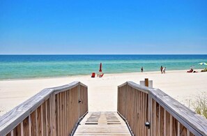 Una passerella di legno che conduce a una spiaggia con acqua turchese cristallina, alcune persone e sedie a sdraio.