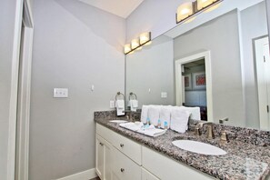 A bathroom with a granite countertop, two sinks, a large mirror, and a door leading to another room. - Our Point of View #3 (Gulf Shores)