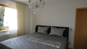 Un dormitorio con una cama grande, una araña, una ventana con persianas y una puerta de madera.