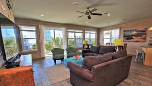 A spacious living room with a brown sofa, green armchairs, and a wooden coffee table. There is a ceiling fan, large windows, and a painting on the wall. - Serendipity Sands (Gulf Shores)