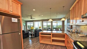A spacious kitchen with wooden cabinets, a refrigerator, and a large island. There is a ceiling fan and pendant lights. - Serendipity Sands (Gulf Shores)
