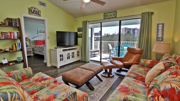 A living room with a sofa, ottoman, and a television. There is a balcony with a chair and a table. - Palmetto 306 (Orange Beach)