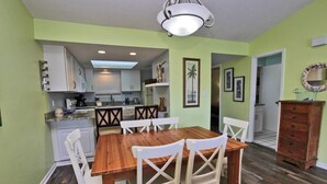 A dining area with a wooden table and white chairs, a kitchen in the background, and a wooden dresser. - Palmetto 306 (Orange Beach)