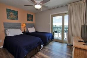 A hotel room with two beds, a ceiling fan, a balcony with curtains, and artwork on the walls.