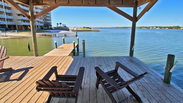 A wooden deck with two Adirondack chairs overlooking a body of water, a pier extending into the water, and a building with balconies in the background. - On Golden Pond 8A West (Gulf Shores)