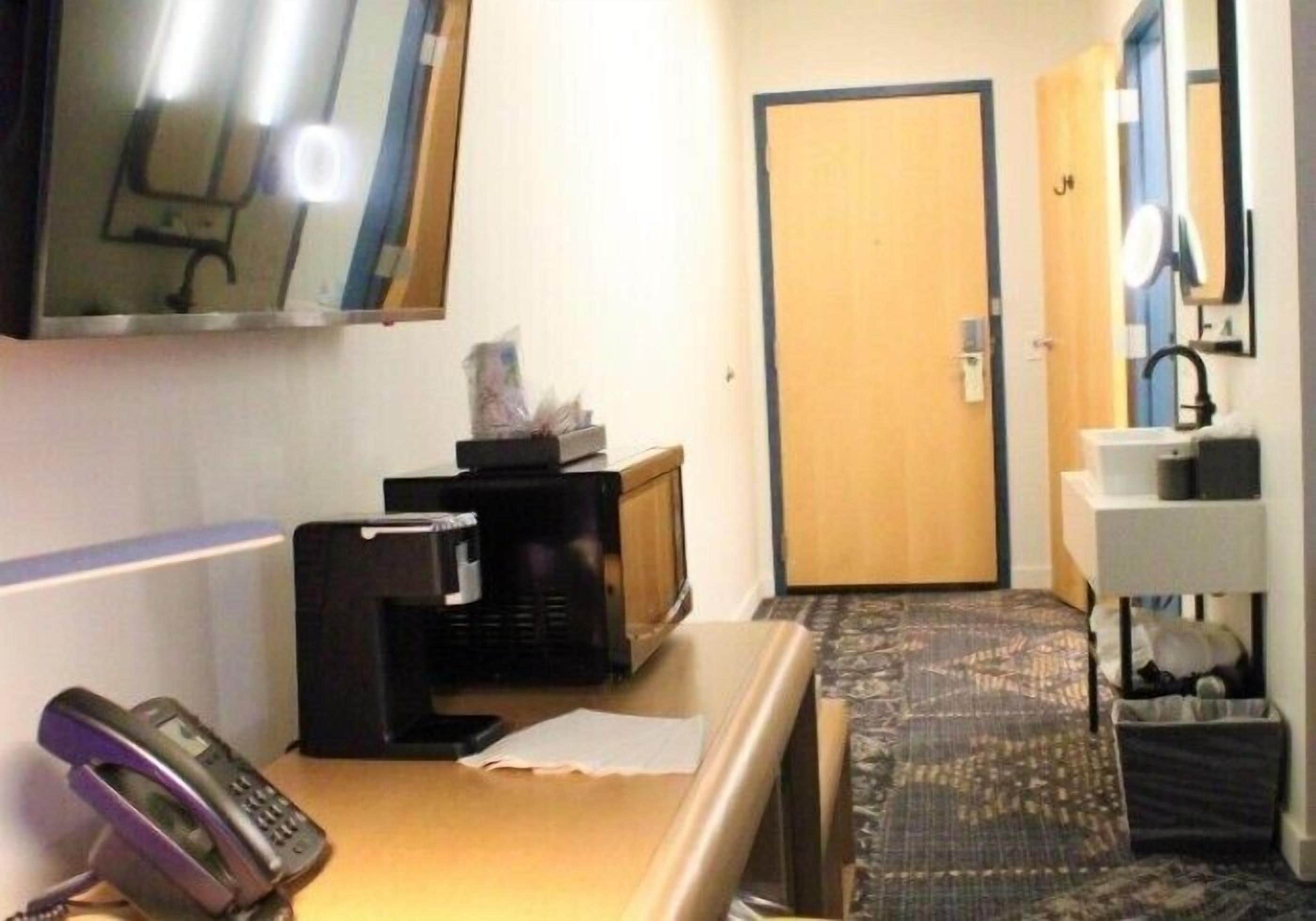 Habitación de hotel con escritorio, teléfono, microondas, televisor pequeño, puerta y espejo.