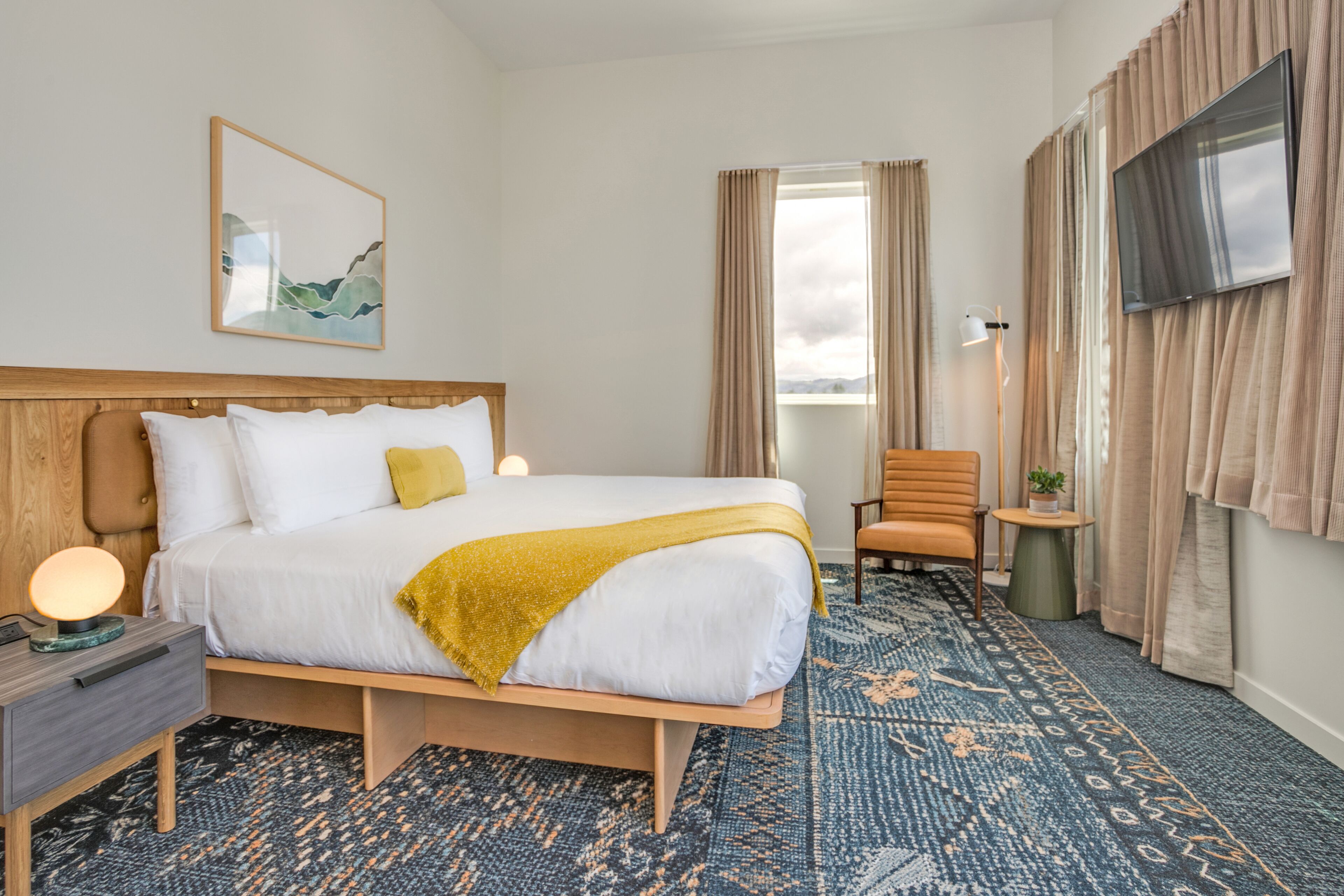A hotel room with a bed, bedside table, lamp, chair, TV, and a window with curtains.