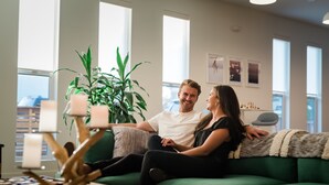 Un couple est assis sur un canapé vert dans une pièce bien éclairée, dotée de grandes fenêtres, d’une plante et d’une lampe décorative.