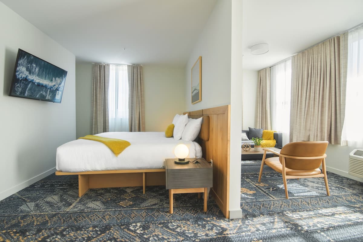 A hotel room with a large bed, a bedside table with a lamp, a chair, a sofa, and a TV mounted on the wall.