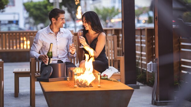 Un couple est assis à une table, devant un petit foyer mobile, avec une bouteille de champagne et des verres.