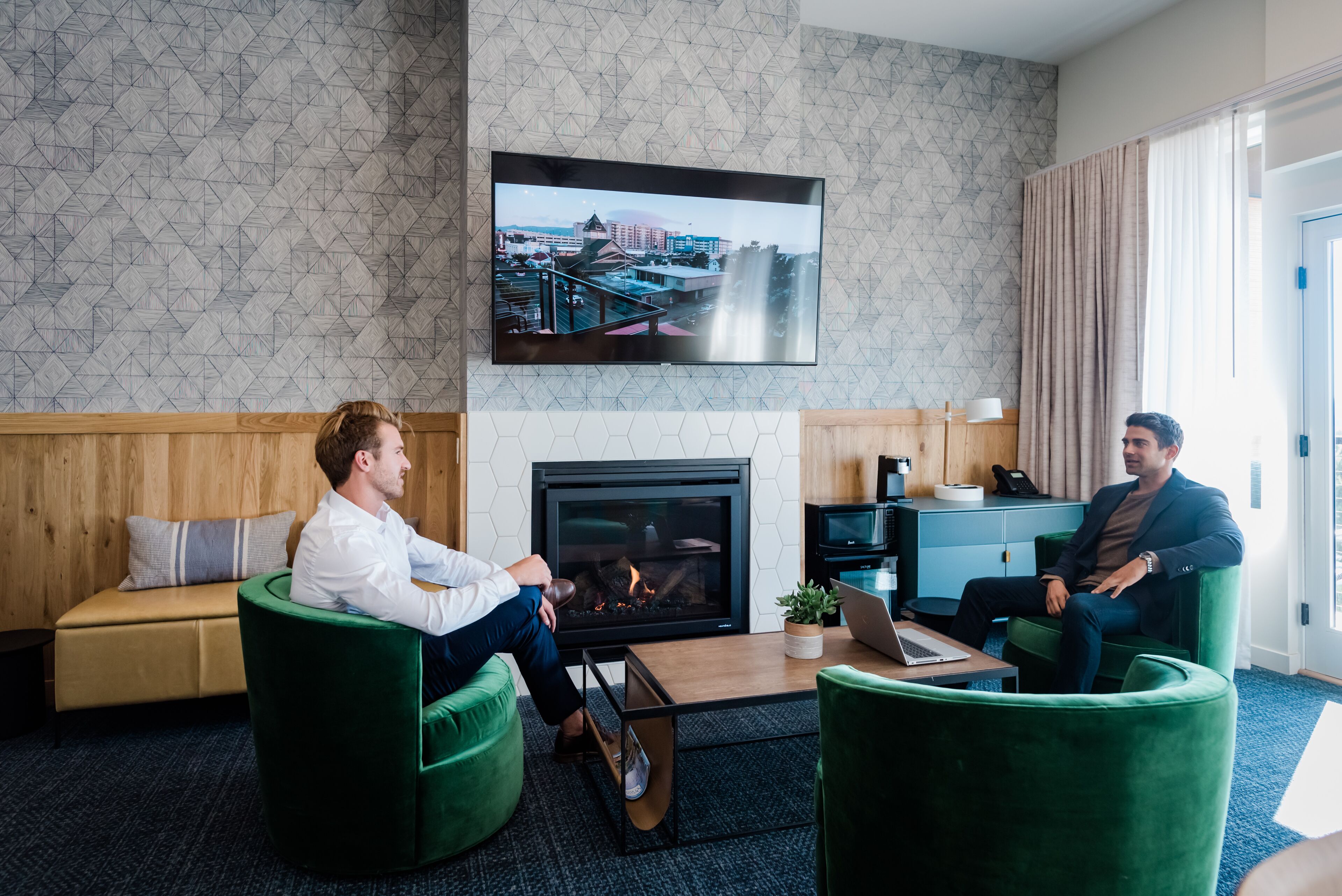 A modern hotel room with a fireplace, a large TV mounted on the wall, and two green armchairs.