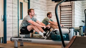 Duas pessoas usando uma máquina de remo em uma academia com espelhos e uma porta de madeira.