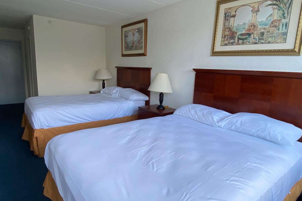A hotel room with two single beds, wooden headboards, white linens, two bedside tables with lamps, and a framed painting on the wall.