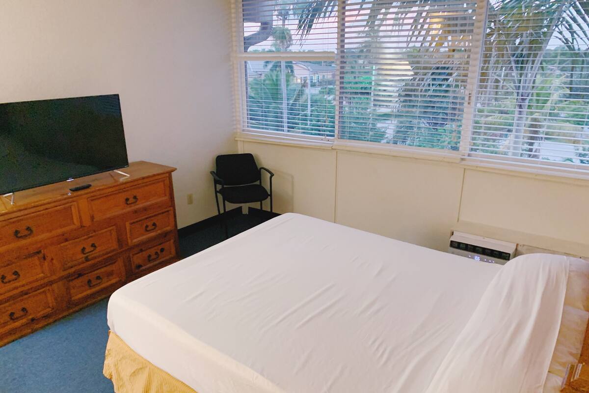 A bedroom with a bed, a wooden dresser, a television, a chair, and a window with blinds.