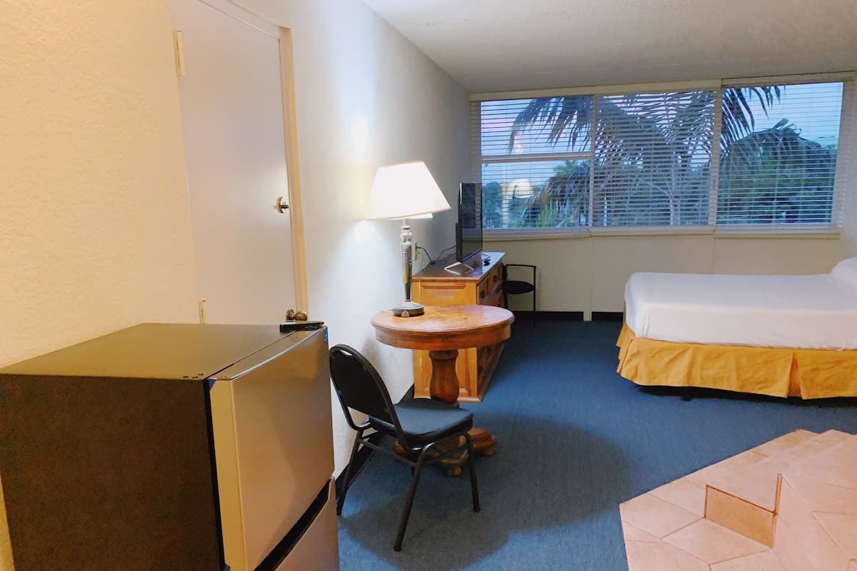A compact hotel room with a bed, a desk with a lamp, a chair, and a refrigerator.