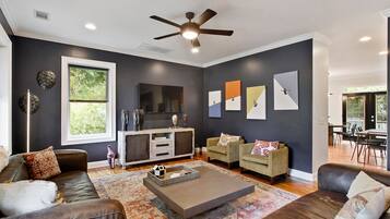 Un salon moderne avec un mur de couleur sombre, un ventilateur de plafond, une grande fenêtre et un coin repas visible à travers une porte ouverte.