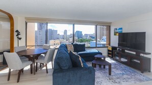 A modern living room with a large TV, a sofa, a dining table, and a city view through large windows. - Discovery Bay, Conveniently Located In Waikiki, near Ala Moana Shopping Center & the beach! (Honolulu)