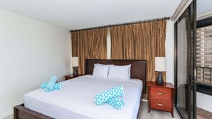 A hotel room with a large bed, two bedside tables with lamps, a sliding glass door, and a view of the cityscape. - Discovery Bay, Conveniently Located In Waikiki, near Ala Moana Shopping Center & the beach! (Honolulu)