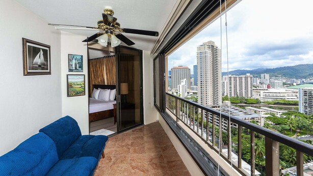 A balcony with a city view, a ceiling fan, and a bed with a view. - Discovery Bay, Conveniently Located In Waikiki, near Ala Moana Shopping Center & the beach! (Honolulu)