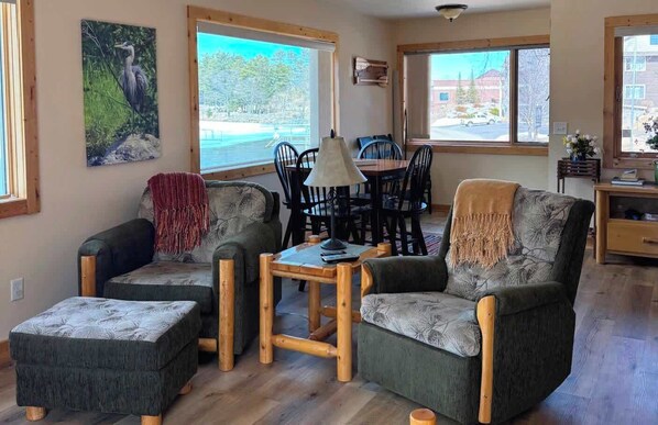 A cozy living room with two sofas, a coffee table, and a dining table with chairs. Large windows offer views of the outdoors.