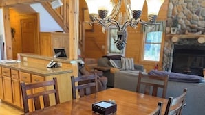 A wooden dining table with chairs, a chandelier, a stone fireplace, and a kitchen area. - Camp Pinemere - Private Northwoods Retreat (Minocqua)