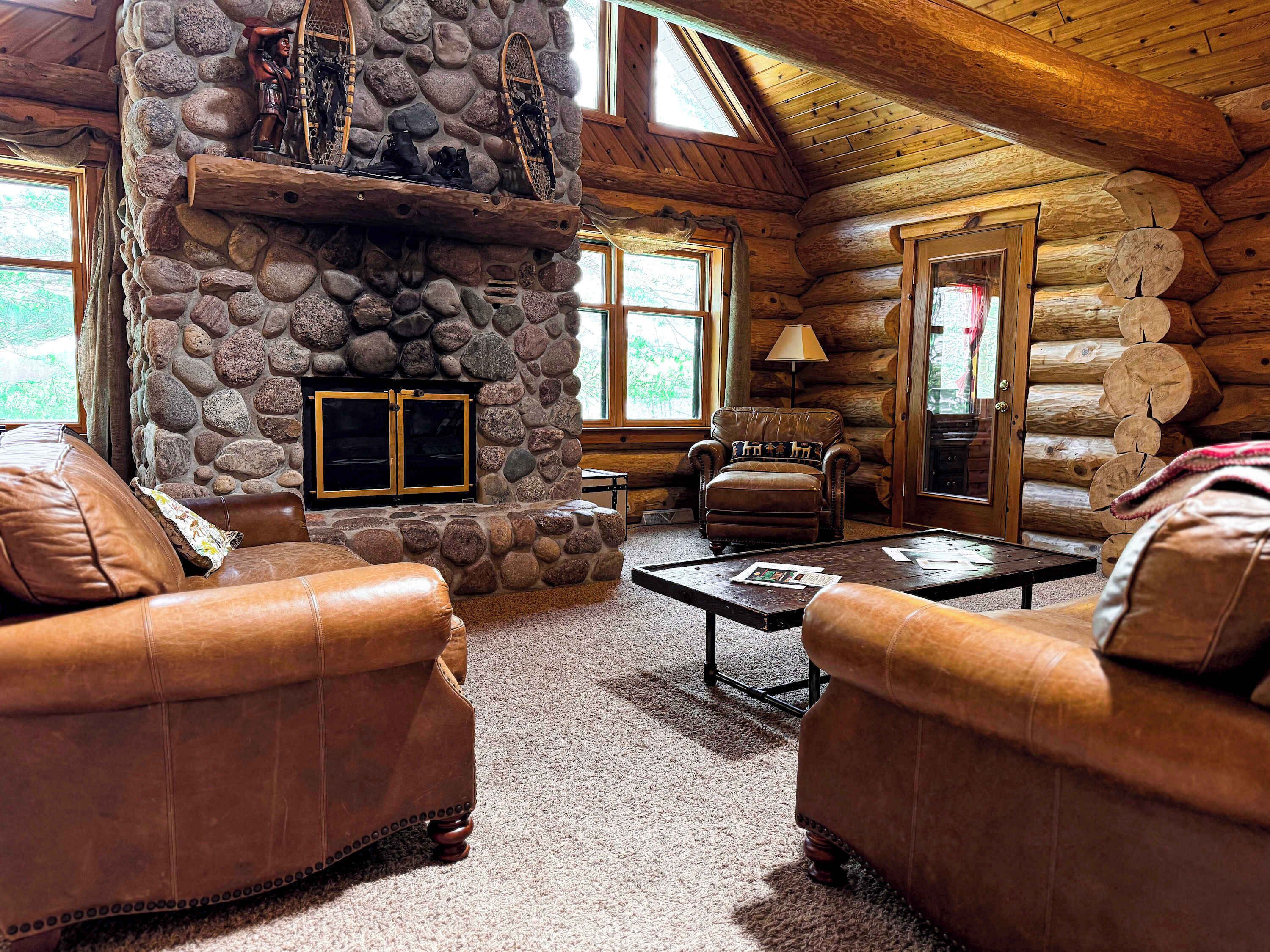A log cabin interior with a stone fireplace, leather sofas, and a wooden ceiling.