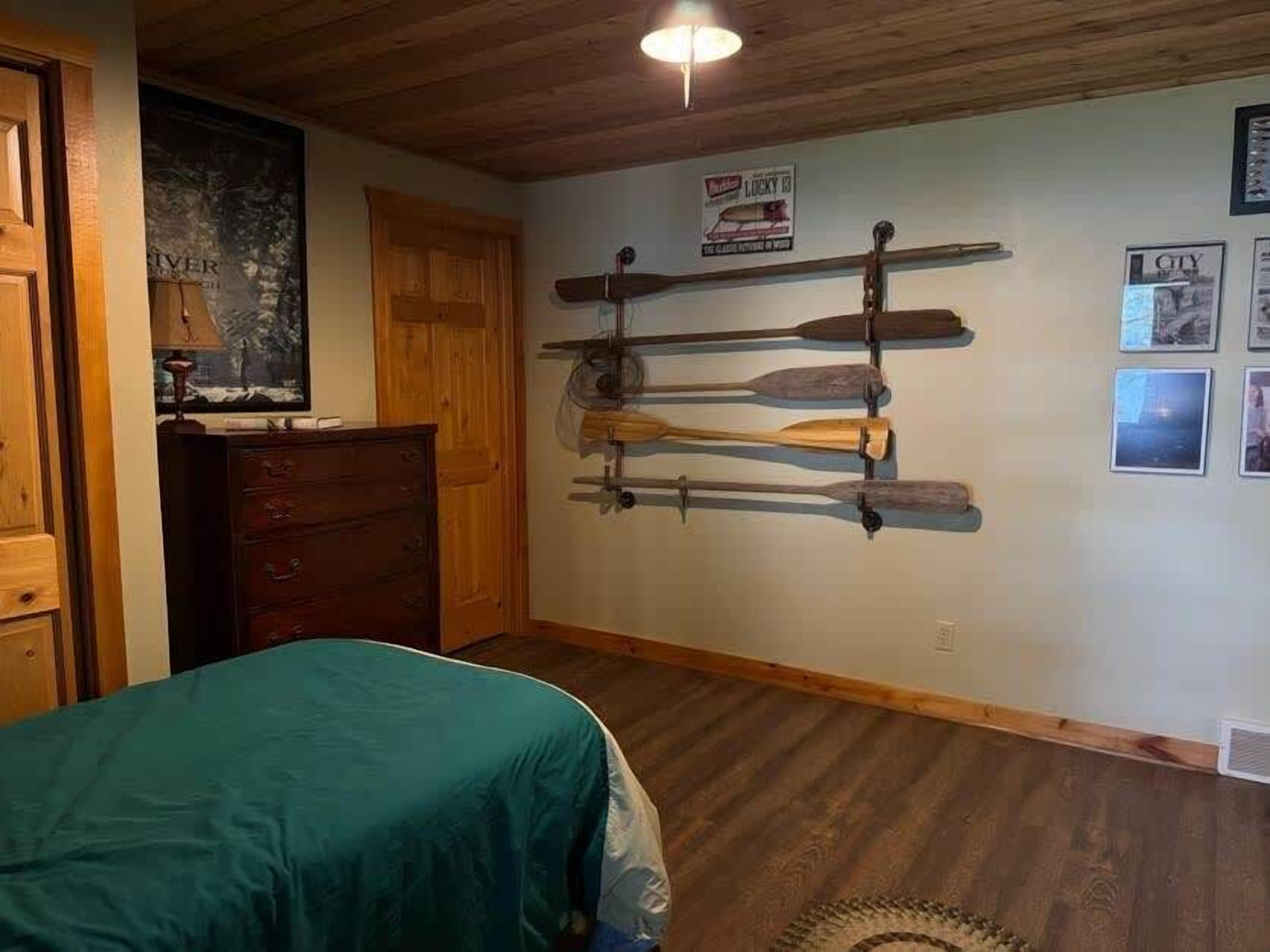 A bedroom with a wooden ceiling, a bed with a teal cover, a wooden dresser, and a wall-mounted paddleboard rack.