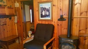 Una habitación acogedora con detalles de madera, una silla negra, una mesita pequeña, un cuadro en la pared y un estufa de leña.
