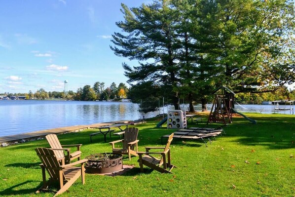 Un parque junto al lago con sillas de madera, una fogata, mesas de picnic y un parque infantil.