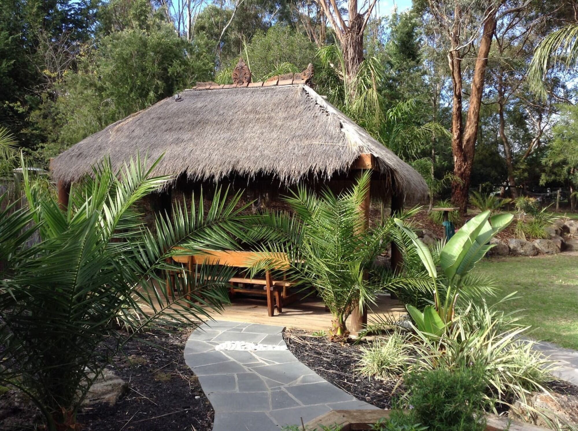 A thatched-roof hut with a bench in front, surrounded by tropical plants and trees.