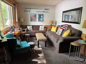 A cozy living room with a sofa, armchairs, a coffee table, and a small side table. There are posters on the wall and a window with curtains.