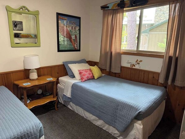 A room with two beds, a wooden nightstand with a lamp, a framed picture on the wall, and a window with curtains.