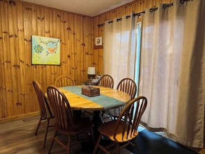 A dining area with a wooden table and chairs, a painting on the wall, and a window with curtains.
