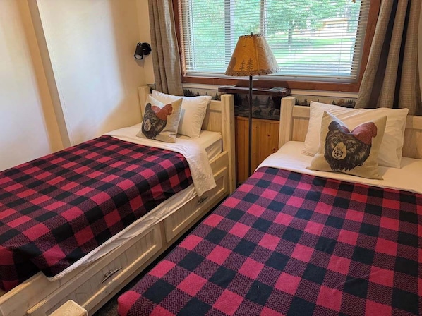 A room with two beds, each with a checkered blanket and a pillow with a dog design. There is a lamp on a wooden nightstand between the beds.