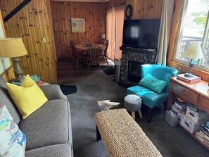 Un salon chaleureux avec un canapé gris, un fauteuil turquoise et un petit pouf. On aperçoit une cheminée au fond, et un téléviseur est fixé au mur.